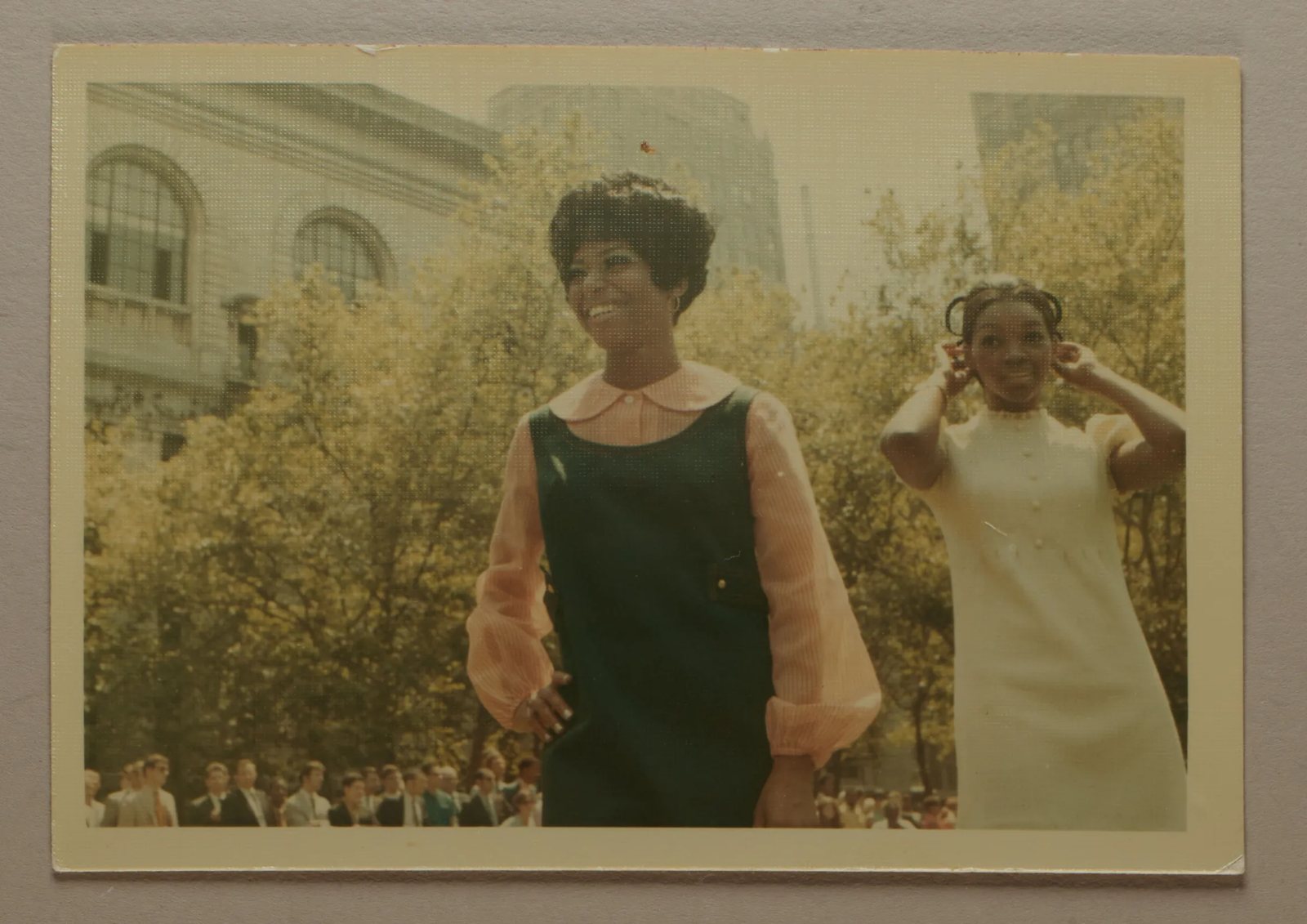 Faded color snapshot of two Black models on an outdoor runway in Bryant Park, one in a green jumper over a pink blouse smiling to the side, the New York Public Library and a crowd of onlookers behind them