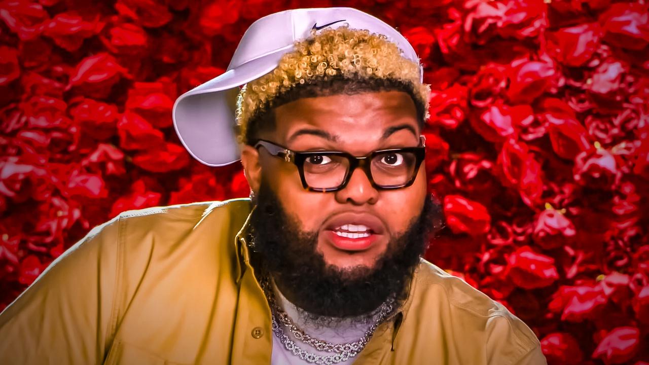 Druski in a backwards white Nike cap, tortoiseshell glasses and a mustard shirt, mid-expression in front of a wall of red roses.
