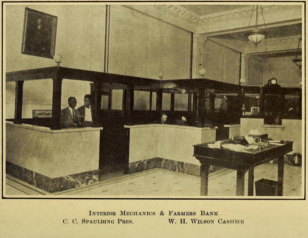 Interior of Mechanics and Farmers Bank in the 1920s: dark wood teller cages, marble base panels, pendant lamps and a framed portrait, with two men standing at the counter.