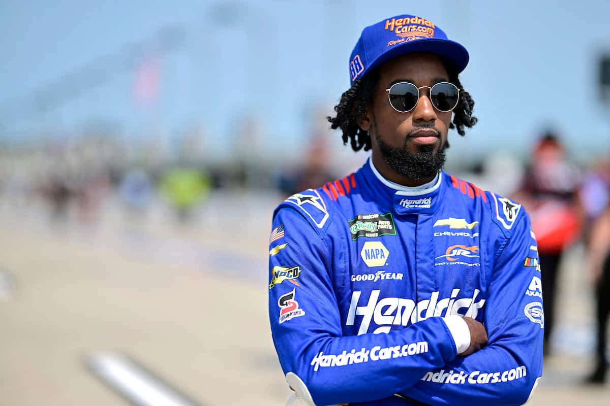 Rajah Caruth on pit road in a blue HendrickCars.com fire suit and cap, arms crossed, wearing round dark sunglasses.